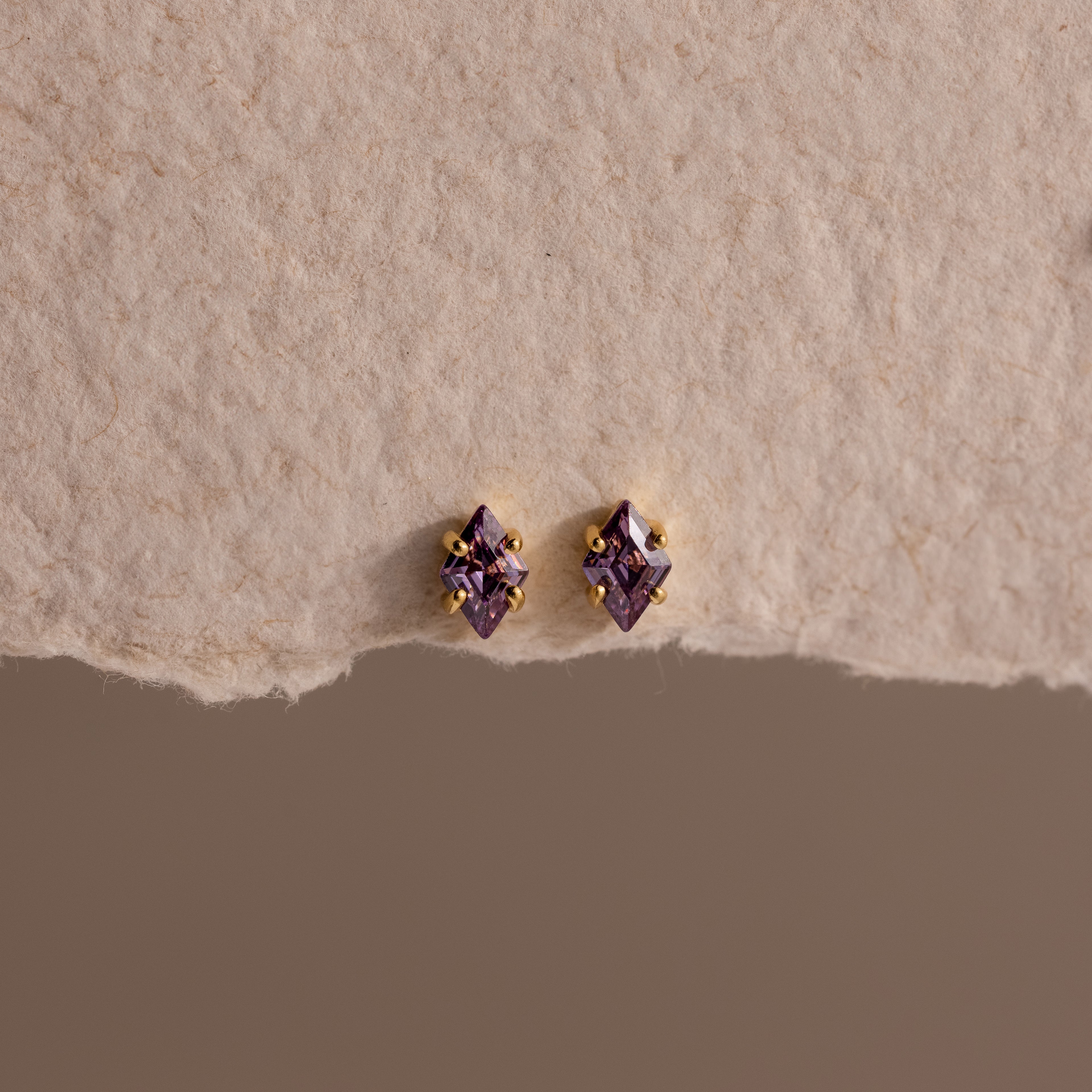 The Rhombus Amethyst Flatback Studs feature small diamond-shaped purple stones with gold accents, adding modern glamour to a chic look. Displayed on a textured beige surface.