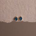 The Rhombus Blue Zircon Flatback Studs, featuring gold accents, offer understated chic with their small blue gemstones, elegantly displayed on a textured beige surface.