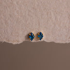 The Rhombus Blue Zircon Flatback Studs, featuring gold accents, offer understated chic with their small blue gemstones, elegantly displayed on a textured beige surface.