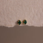 The Rhombus Emerald Flatback Studs are small gold earrings with green gemstone studs, embodying understated chic and presented on textured beige paper.
