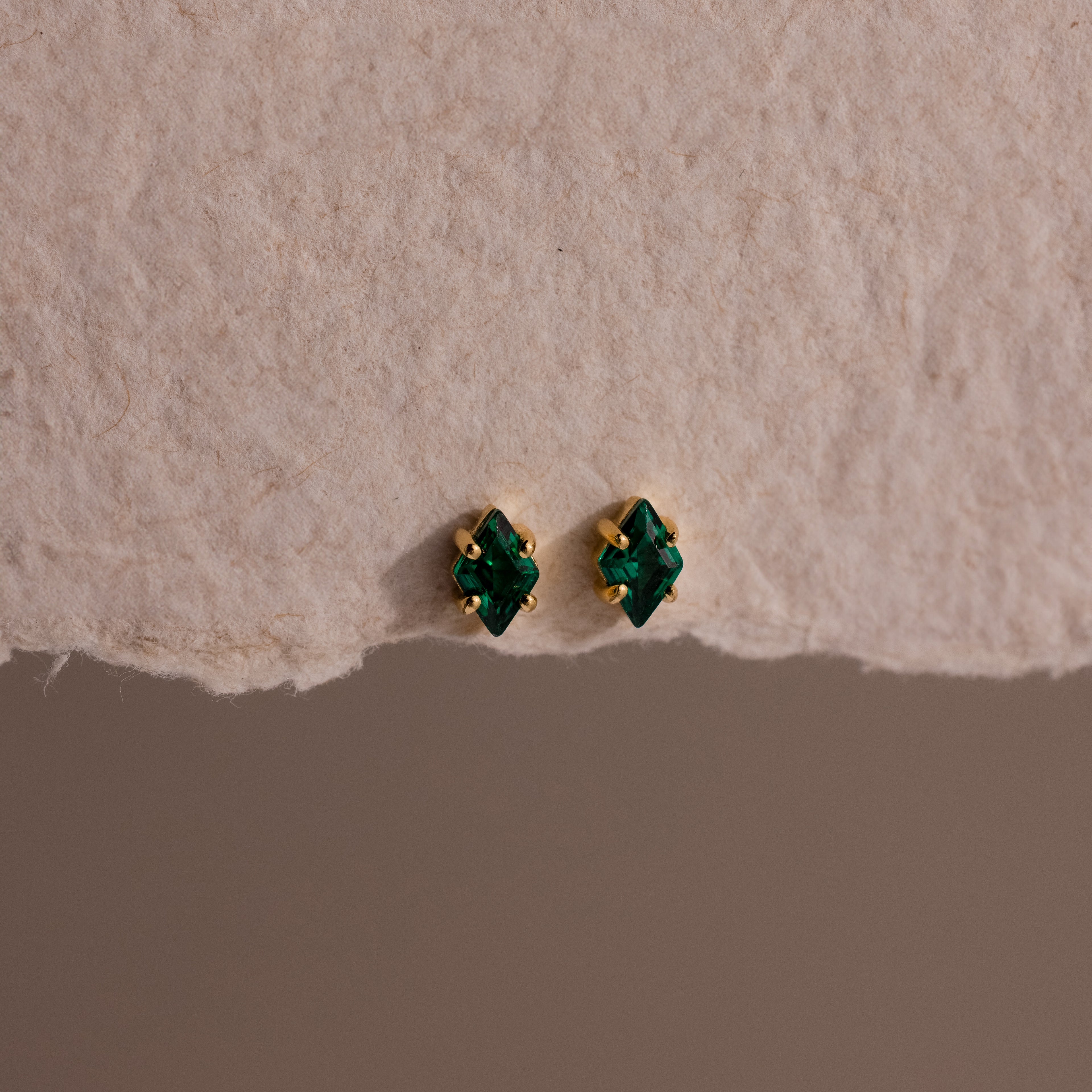 The Rhombus Emerald Flatback Studs are small gold earrings with green gemstone studs, embodying understated chic and presented on textured beige paper.