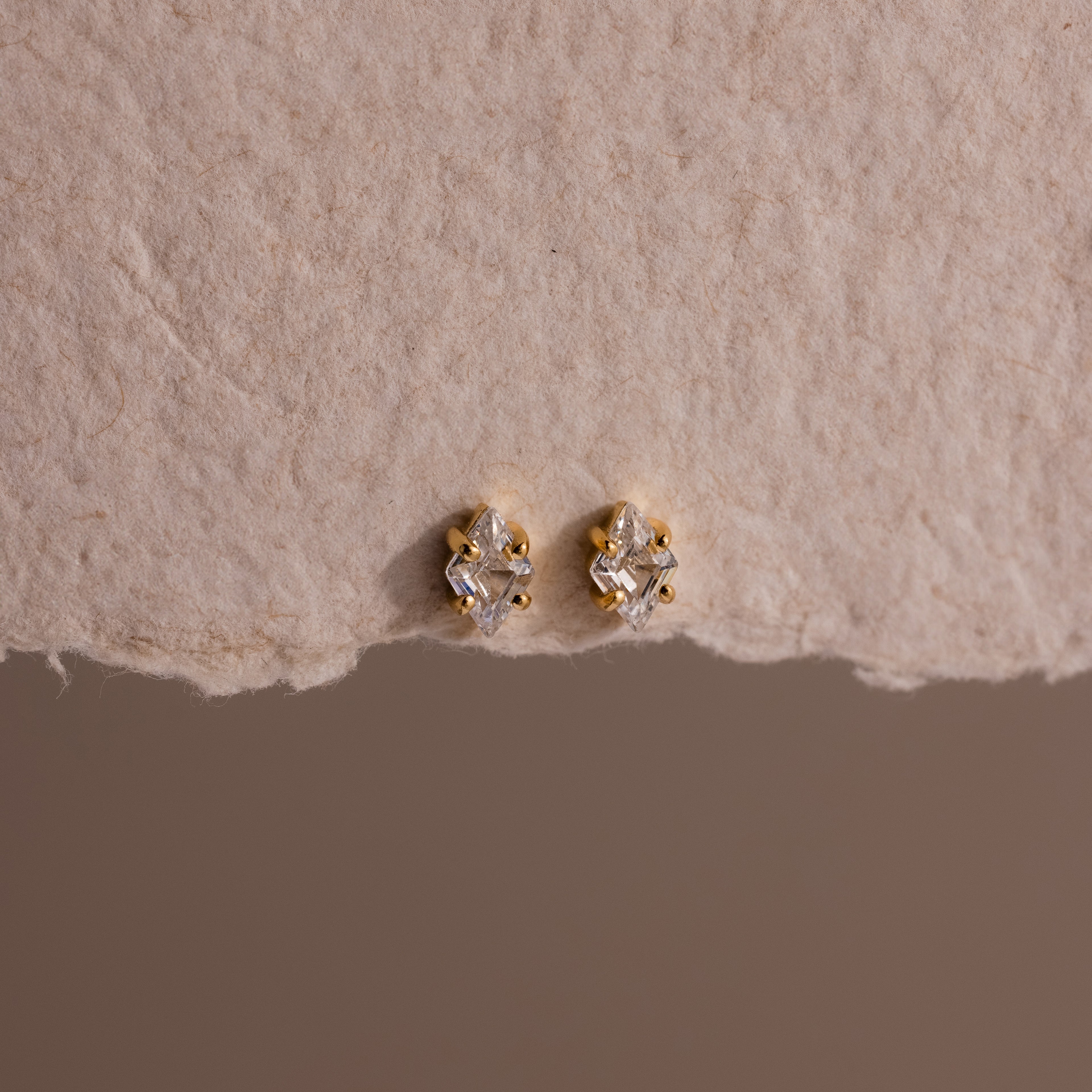 Rhombus Diamond Flatback Studs and other gold and diamond earrings are displayed on textured beige paper, creating an understated chic jewelry look.