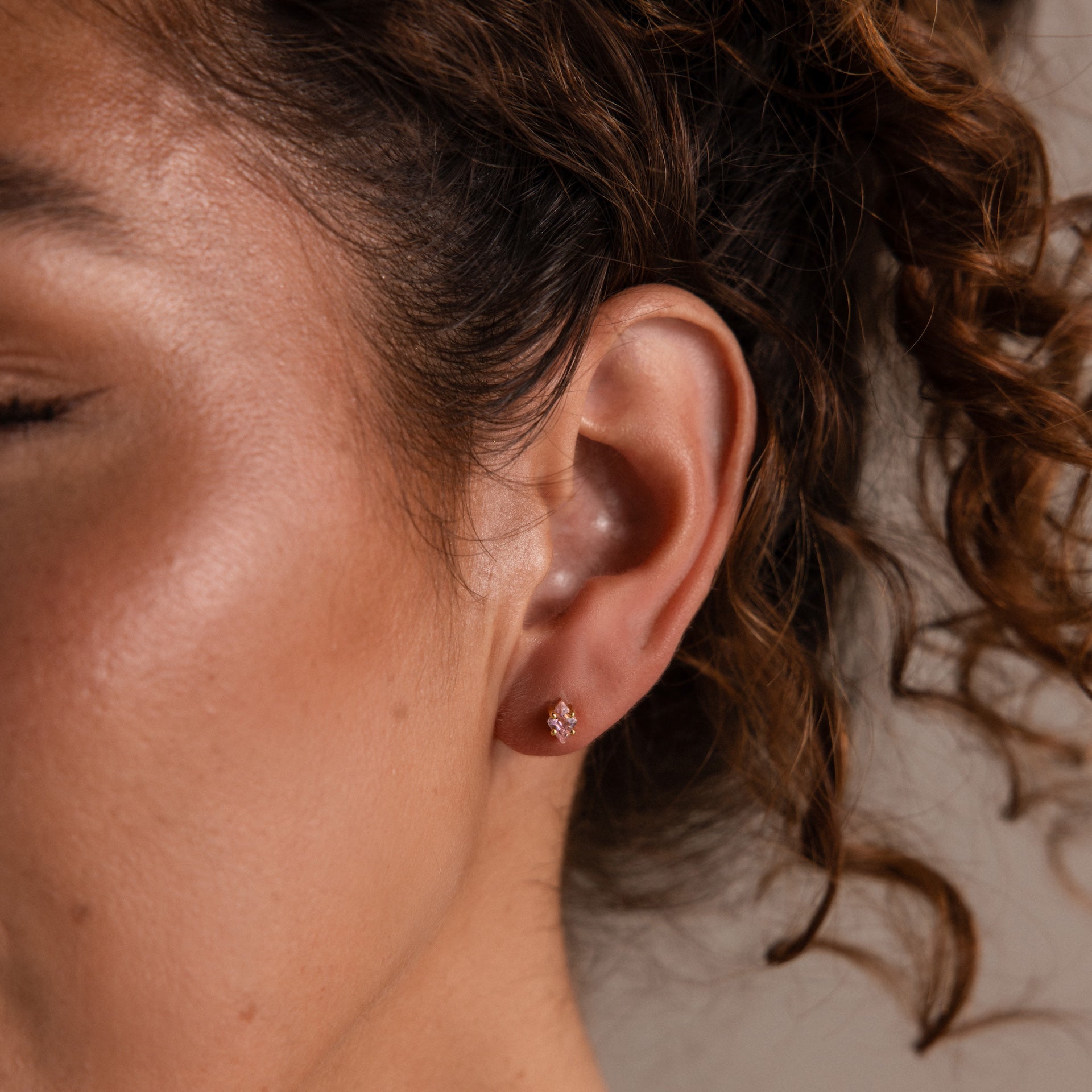 A woman's ear adorned with Rhombus Topaz Flatback Studs sparkles with modern glamour, while her curly hair softly frames her face.