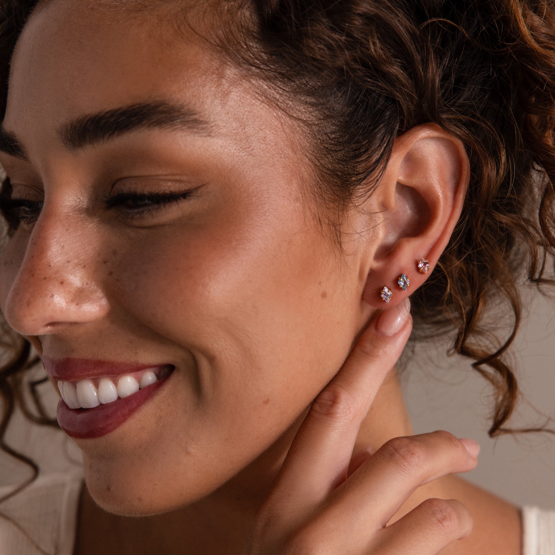 A smiling woman with curly hair showcases Rhombus Birthstone Flatback Studs—understated chic jewelry that captures the modern glamour earrings trend.
