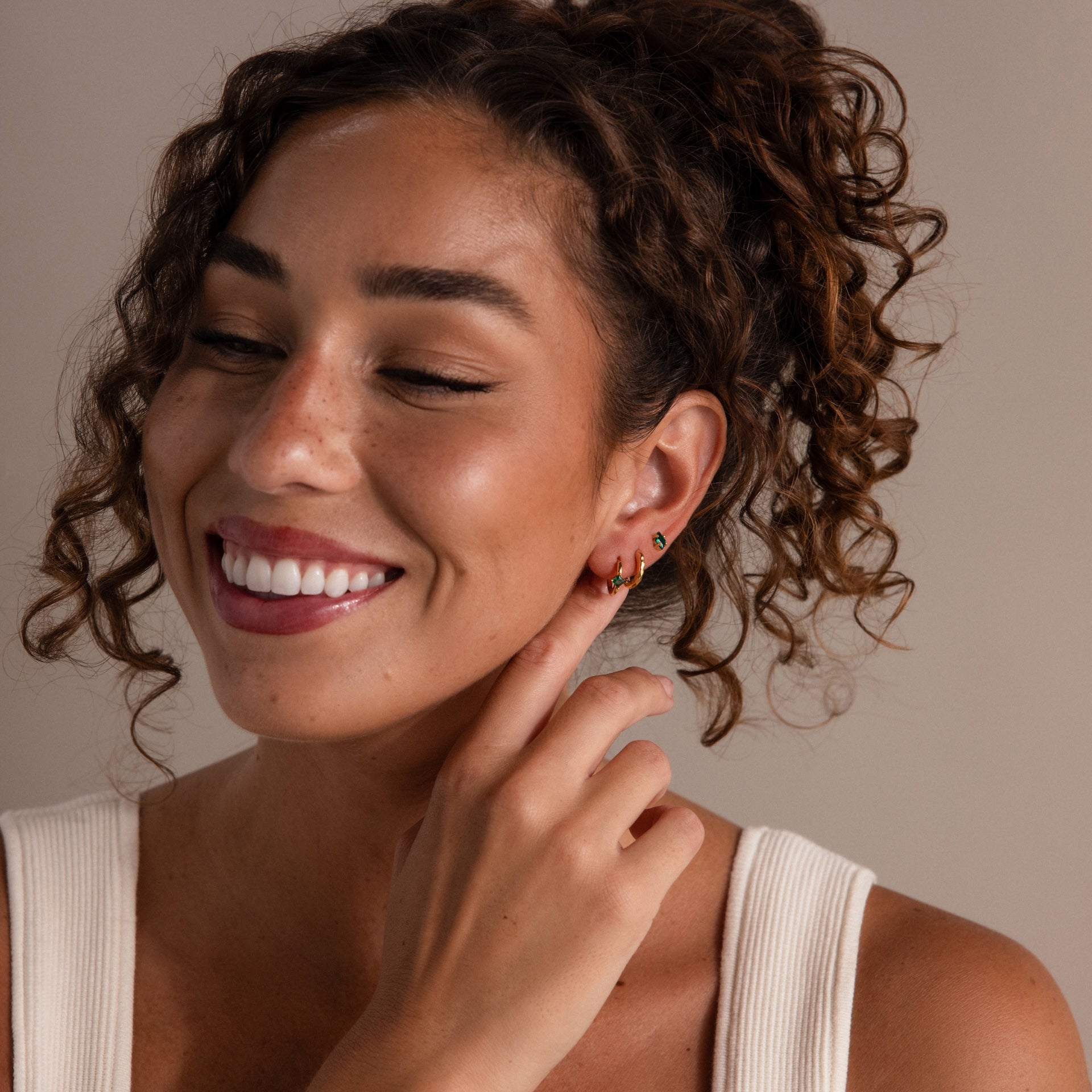 A smiling woman with curly hair touches her Rhombus Topaz Flatback Studs, adding a hint of modern glamour to her look as she wears a white sleeveless top.