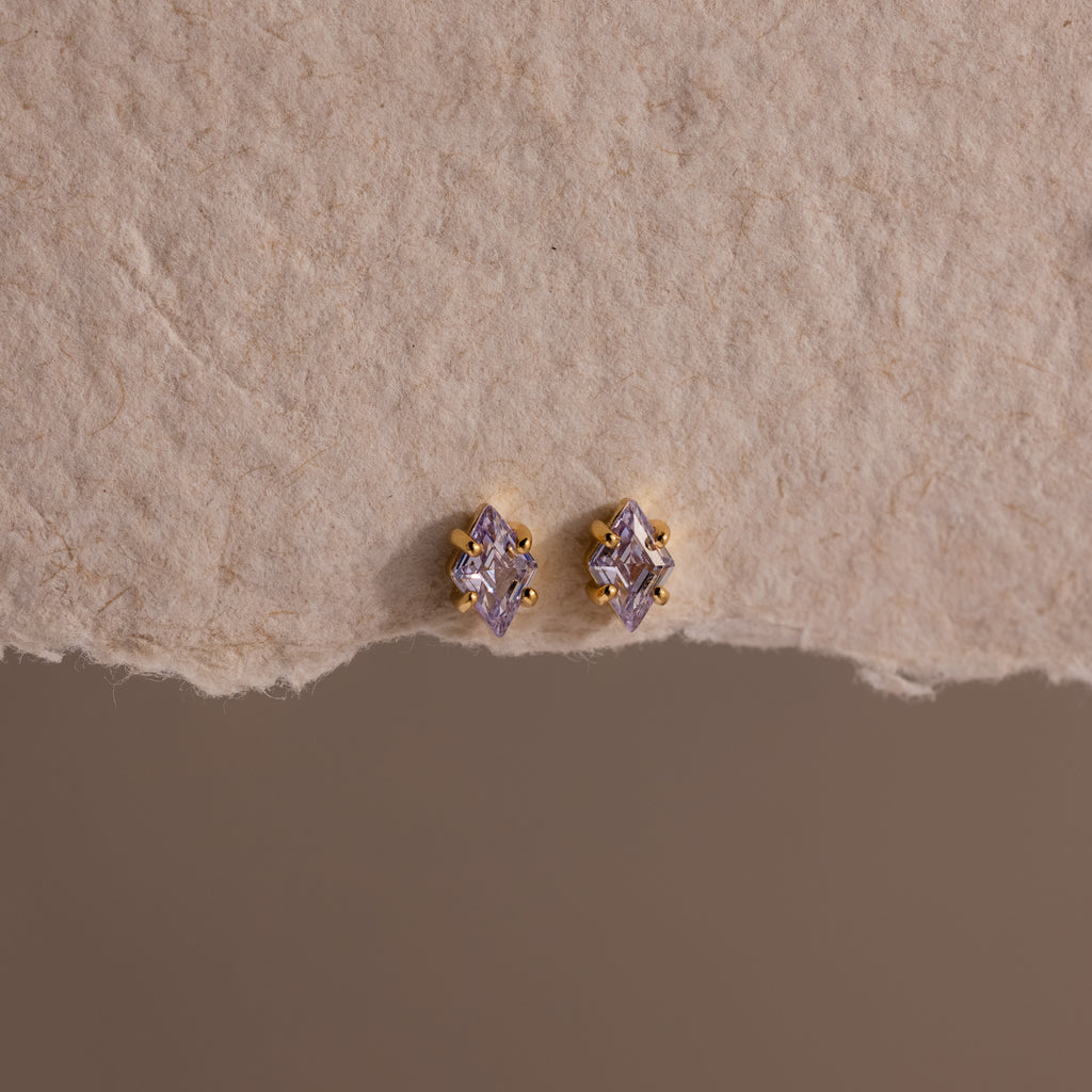 The Rhombus Birthstone Flatback Studs in Alexandrite (June) display modern glamour with their purple marquise-cut stones, elegantly set on a textured beige surface.