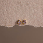 The Rhombus Birthstone Flatback Studs in Alexandrite (June) display modern glamour with their purple marquise-cut stones, elegantly set on a textured beige surface.