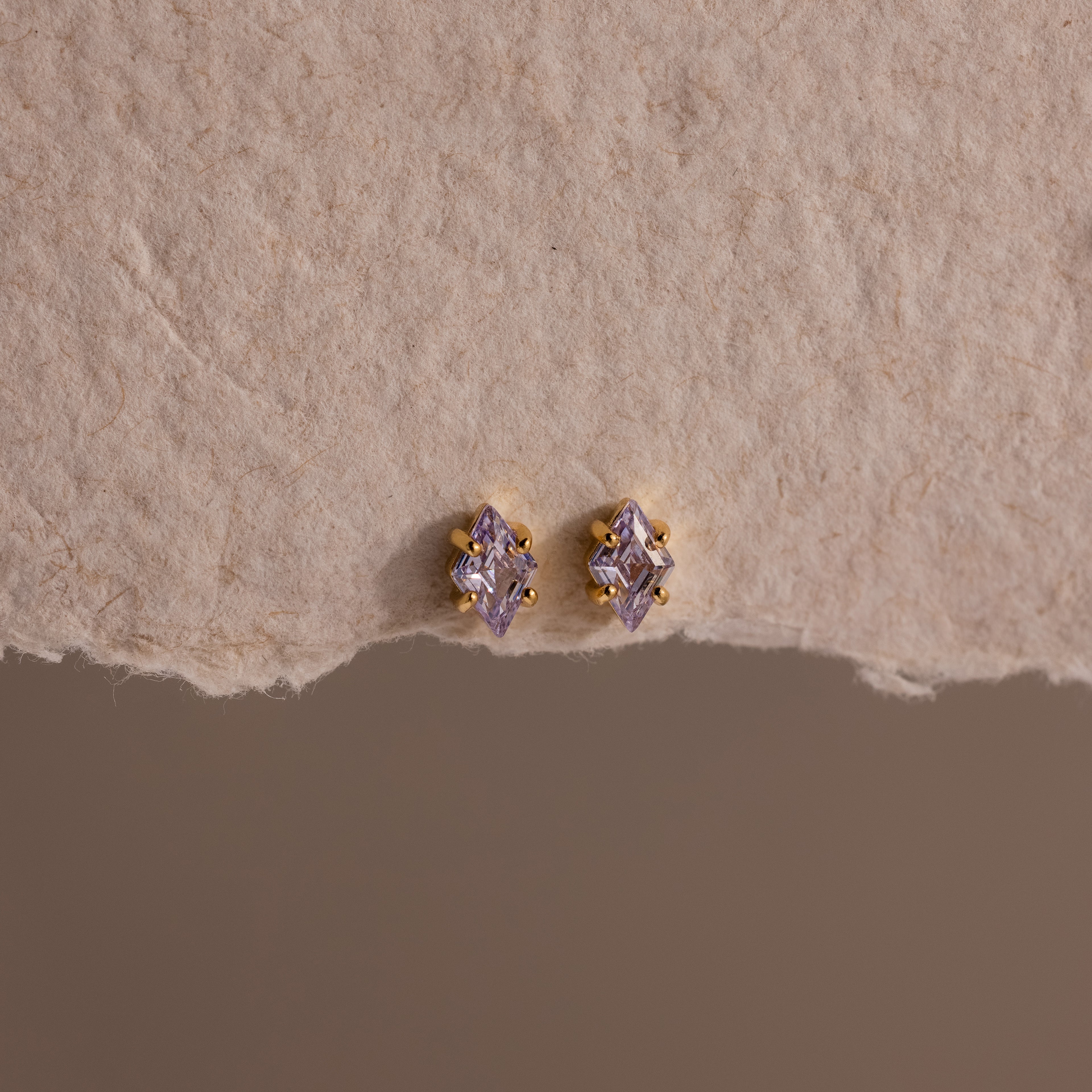 The Rhombus Birthstone Flatback Studs in Alexandrite (June) display modern glamour with their purple marquise-cut stones, elegantly set on a textured beige surface.