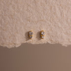 The Tiny Duo Diamond Flatback Studs are small gold earrings featuring two clear gemstones each, ideal for ear stacking or as subtle diamond accents, shown on a textured beige surface.