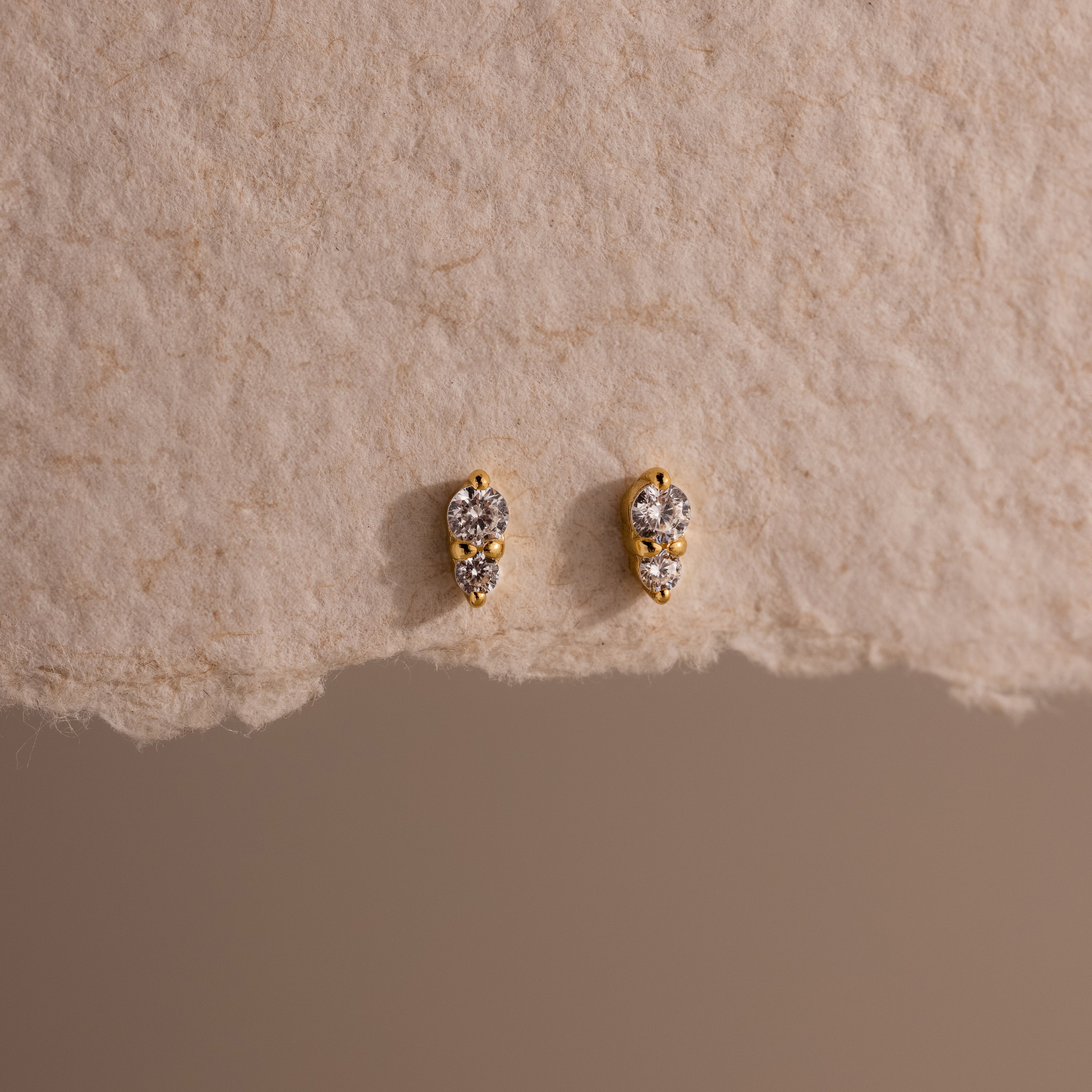 The Tiny Duo Diamond Flatback Studs are small gold earrings featuring two clear gemstones each, ideal for ear stacking or as subtle diamond accents, shown on a textured beige surface.