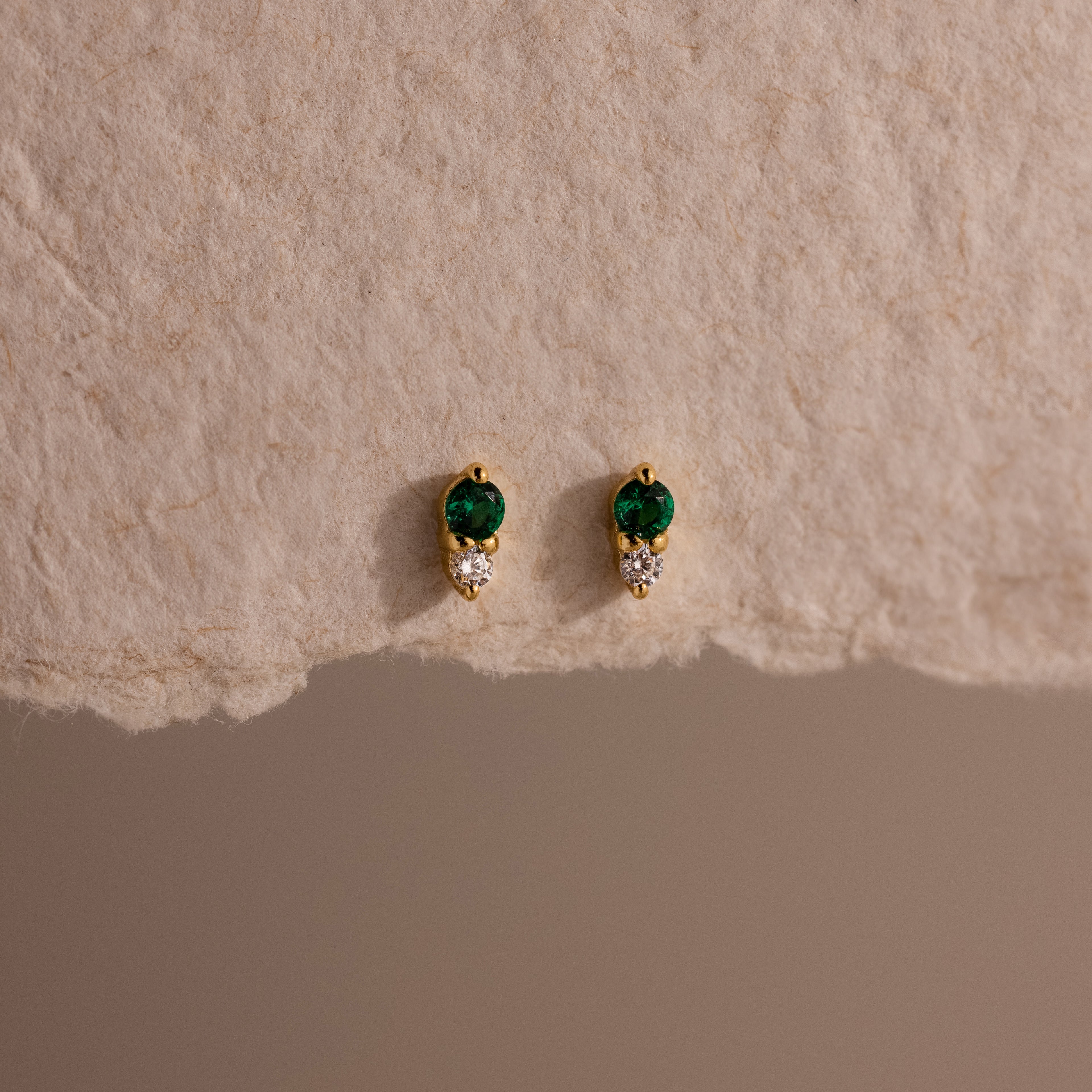 Tiny Duo Emerald Flatback Studs with green and clear stones, perfect for ear stacking, displayed on a textured beige surface.