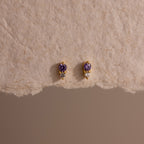 Tiny Duo Amethyst Flatback Studs—gold earrings adorned with small purple gemstones and clear crystals—are displayed on a textured beige surface.