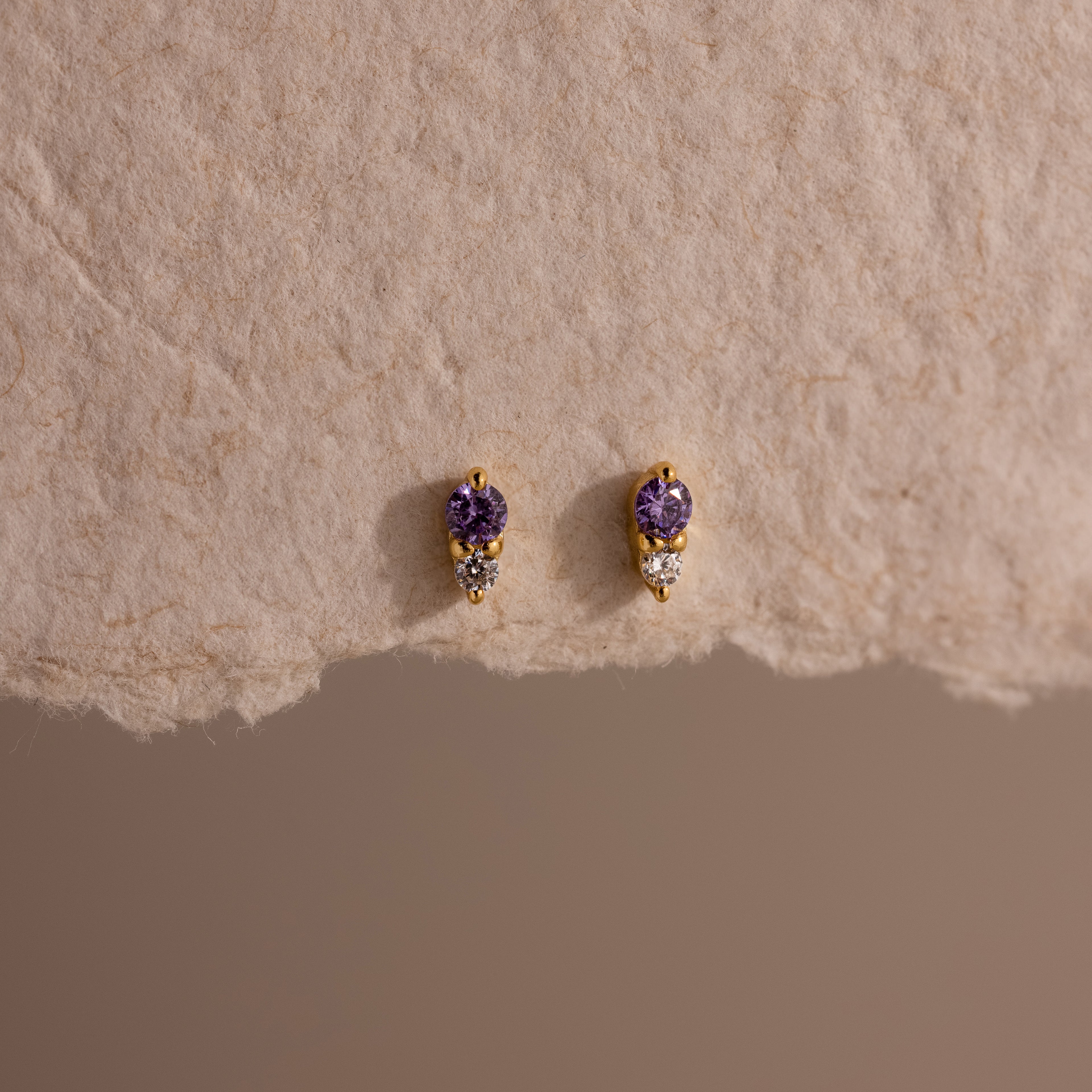 Tiny Duo Amethyst Flatback Studs—gold earrings adorned with small purple gemstones and clear crystals—are displayed on a textured beige surface.
