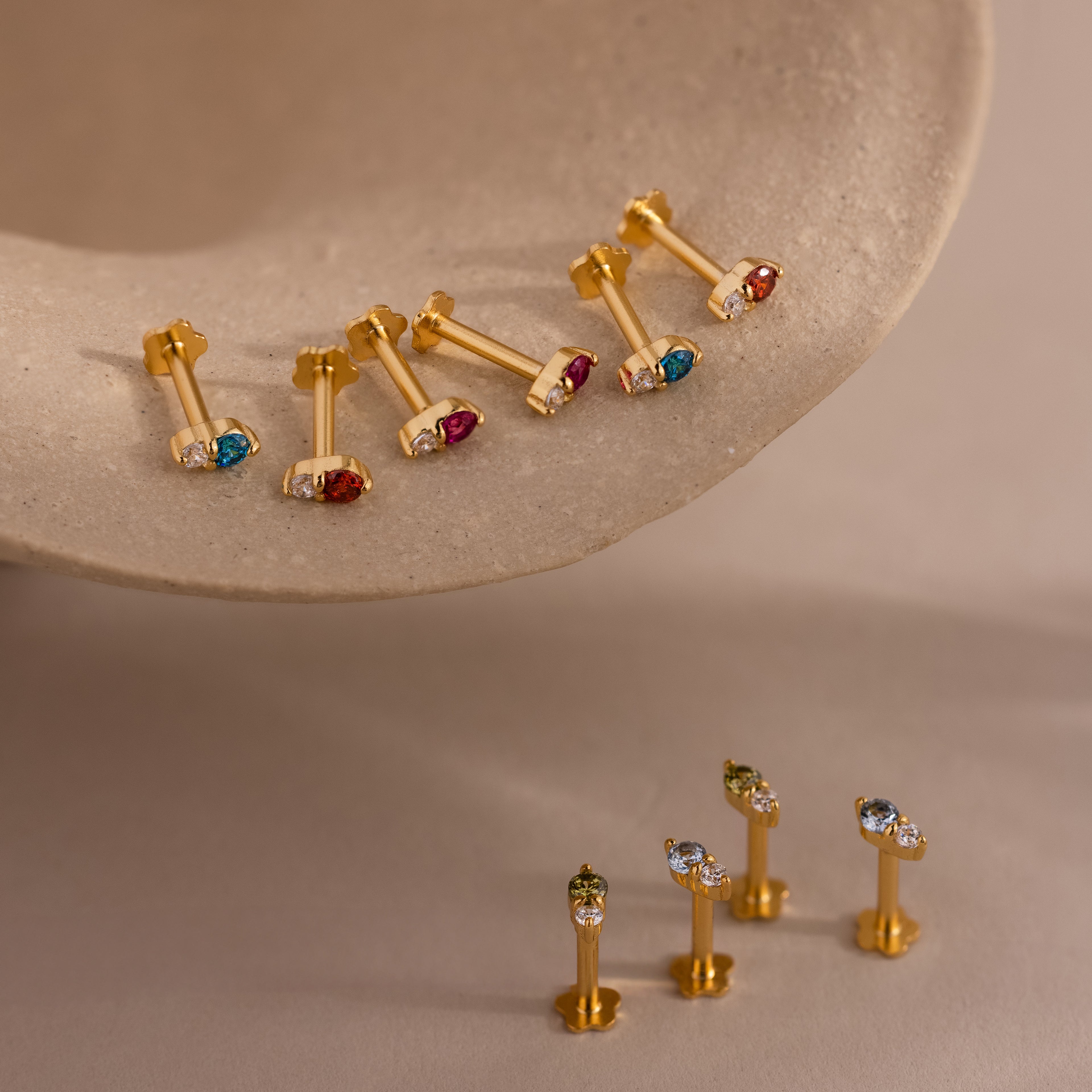 Six gold earrings with colorful gemstones on a beige surface, and four more upright below—perfect for showcasing vibrant ear stacks or highlighting Tiny Duo Blue Zircon Flatback Studs as bold birthstone accents.
