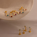 Our Tiny Duo Birthstone Flatback Studs, gold stud earrings with colorful gemstones, are showcased on a beige stone surface set against a neutral background.