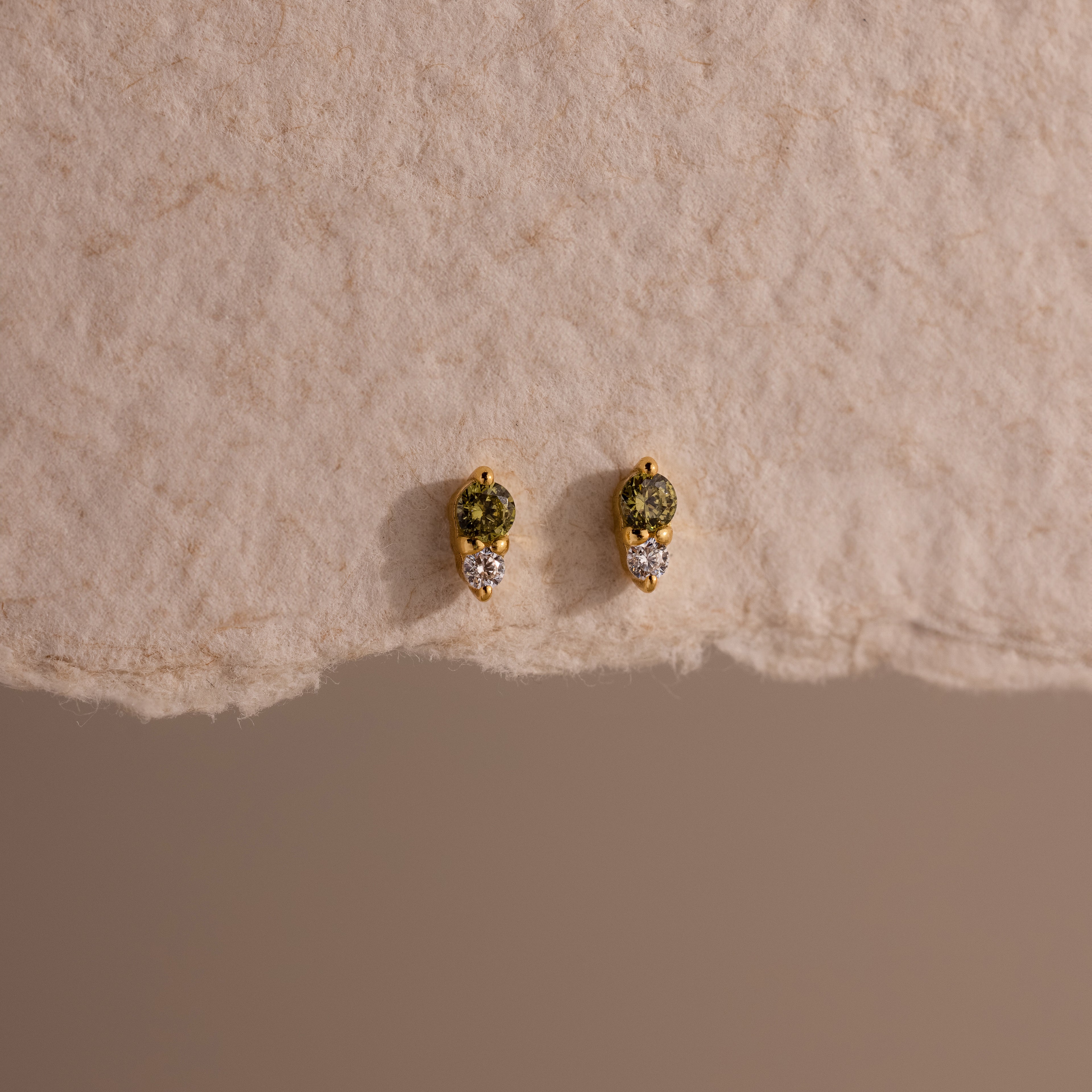 Tiny Duo Peridot Flatback Studs—gold earrings featuring green peridot stones and diamond accents, shown on a textured beige surface—perfect for adding an elegant touch to your ear stack.
