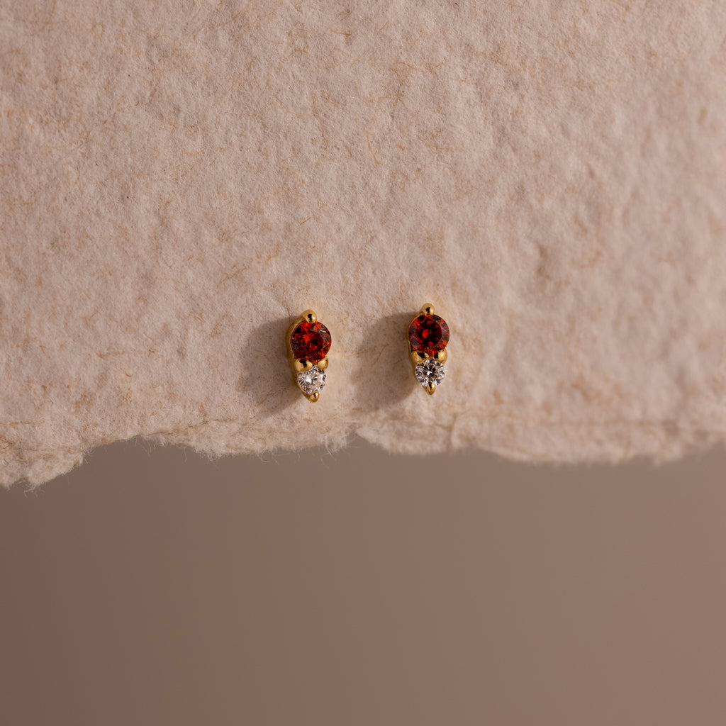 The Tiny Duo Garnet Flatback Studs are small gold earrings with red and white stones, perfect for an ear stack or as elegant birthstone studs, shown on a textured beige surface.