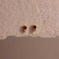 The Tiny Duo Garnet Flatback Studs are small gold earrings with red and white stones, perfect for an ear stack or as elegant birthstone studs, shown on a textured beige surface.