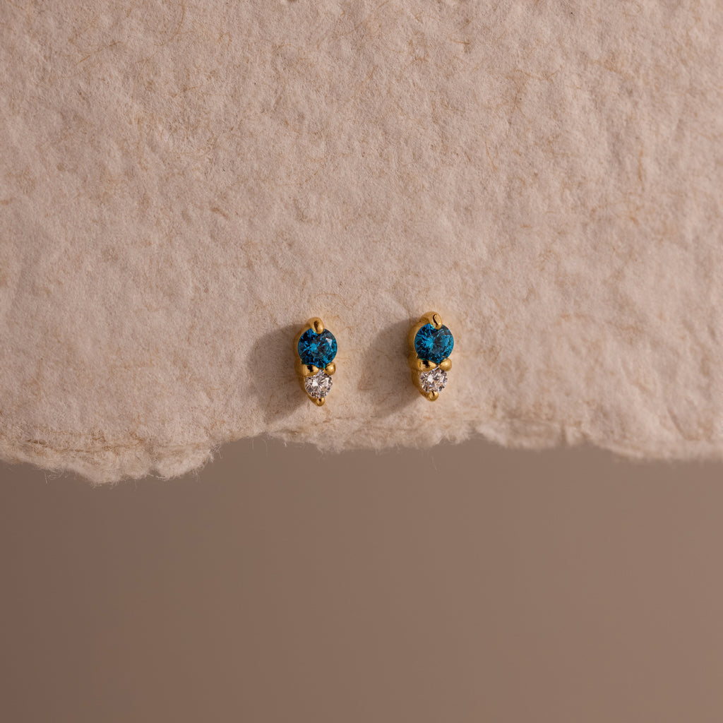 Tiny Duo Blue Zircon Flatback Studs, featuring blue and clear stones, are perfect for an ear stack and shown on a textured beige surface.