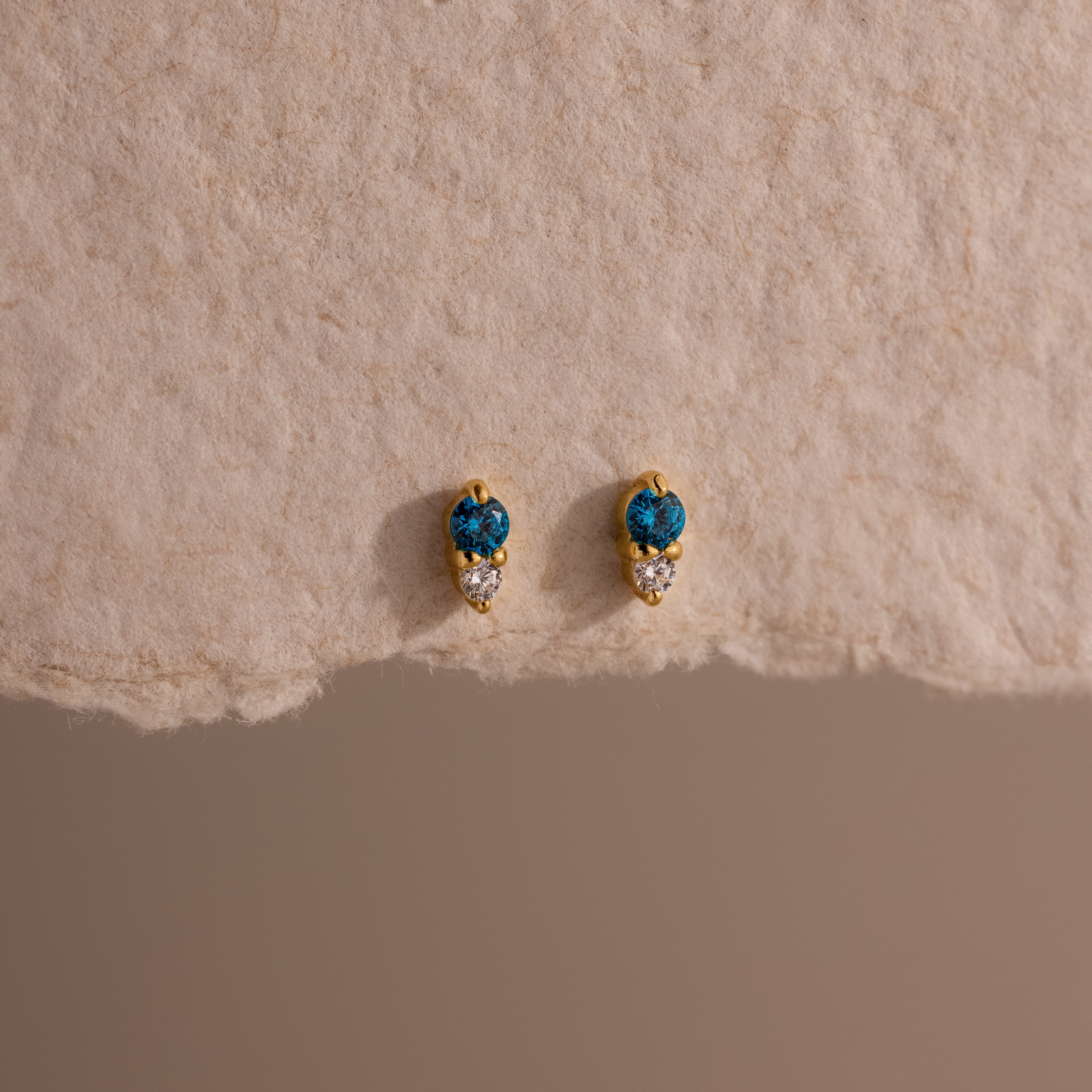 Tiny Duo Blue Zircon Flatback Studs, featuring blue and clear stones, are perfect for an ear stack and shown on a textured beige surface.
