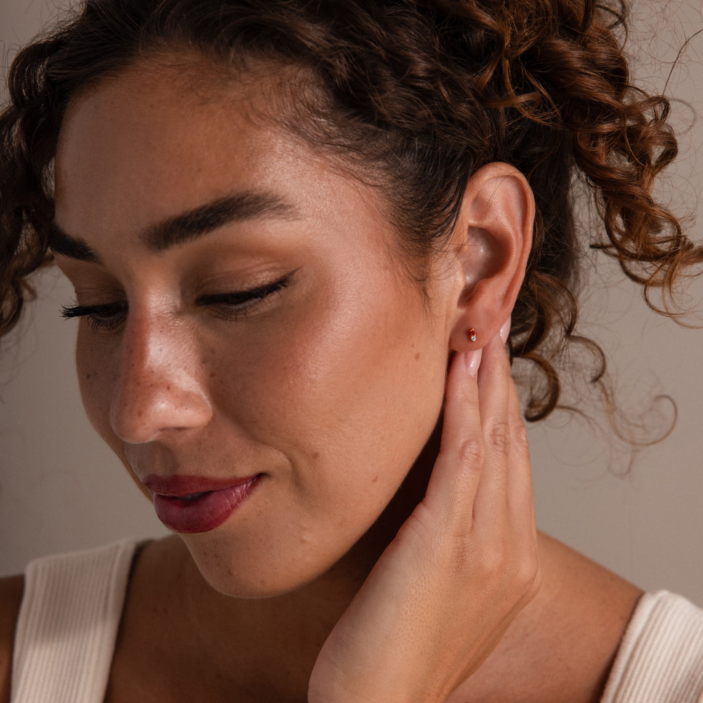 Woman with curly hair touches her ear and smiles softly, wearing a white top and Tiny Duo Birthstone Flatback Studs that add a subtle pop of color.