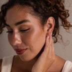 Woman with curly hair touches her ear and smiles softly, wearing a white top and Tiny Duo Birthstone Flatback Studs that add a subtle pop of color.
