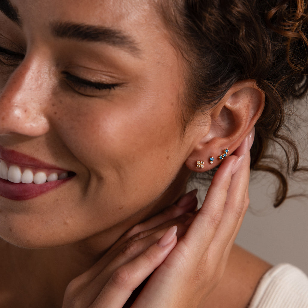 Smiling woman with curly hair touches her ear, wearing Tiny Duo Birthstone Flatback Studs that add a touch of sparkle to her look.