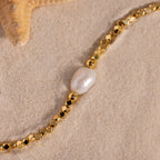 Close-up of a gold beaded bracelet with a single organic freshwater pearl centerpiece, styled on sand next to a starfish.