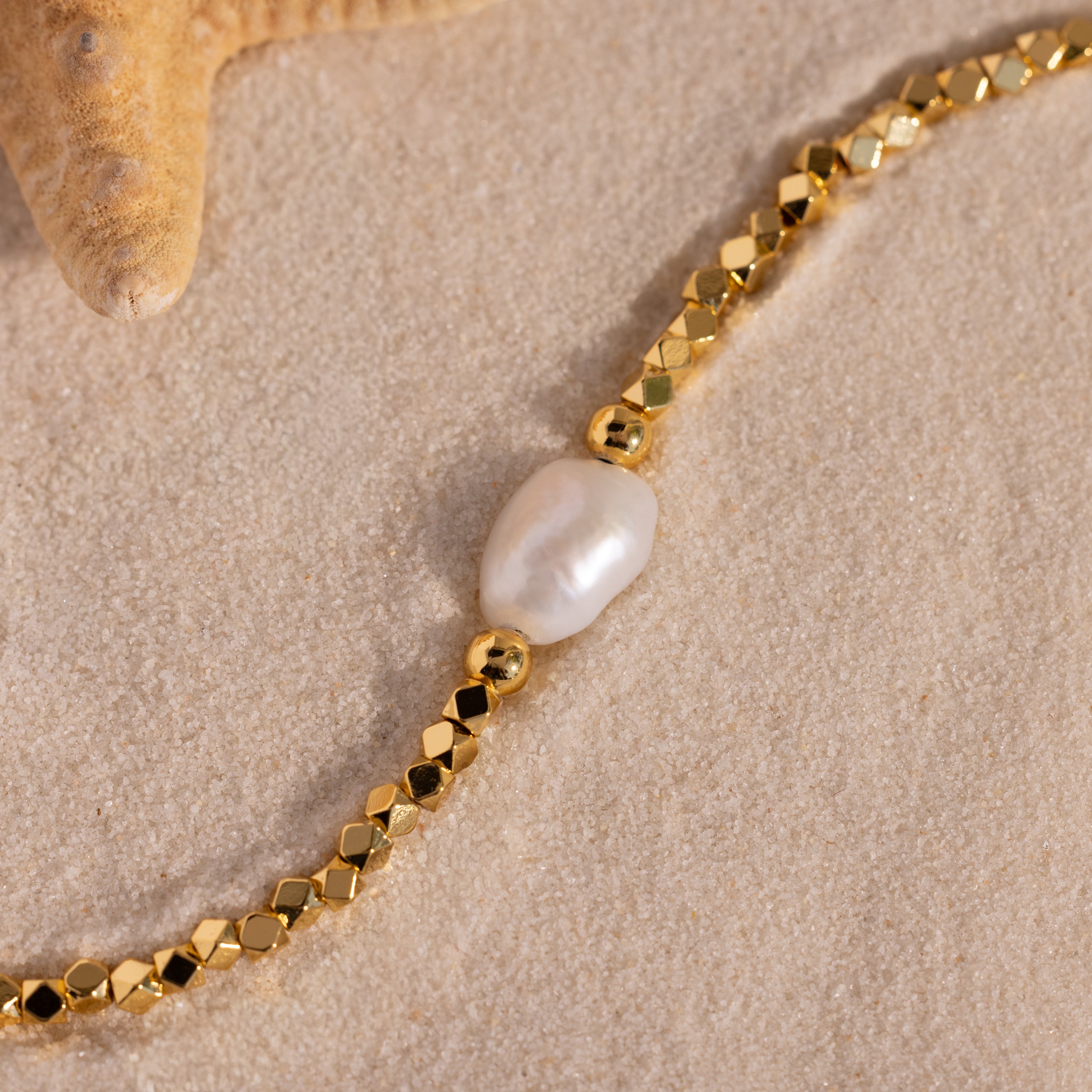 Close-up of a gold beaded bracelet with a single organic freshwater pearl centerpiece, styled on sand next to a starfish.
