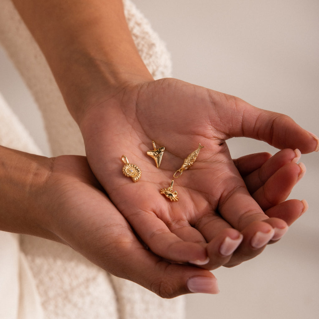 Hand holding five small gold charms, including a pavé seashell charm, a shark tooth, a fish, a lion head, and a sea creature charm, all finely detailed and glinting under soft light.