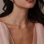 A woman wears a pink sheer top with layered gold necklaces, including the Mirror Chain Lariat Necklace, and small daisy earrings, shown from the shoulders up.