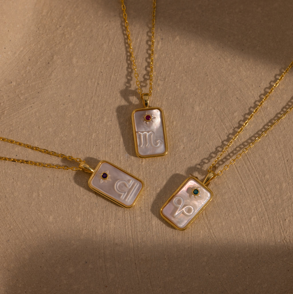 A Pearl Zodiac Birthstone Necklace in Rose Gold, featuring a rectangular zodiac pendant and delicate birthstone, is displayed as personalized jewelry on a neutral textured background.
