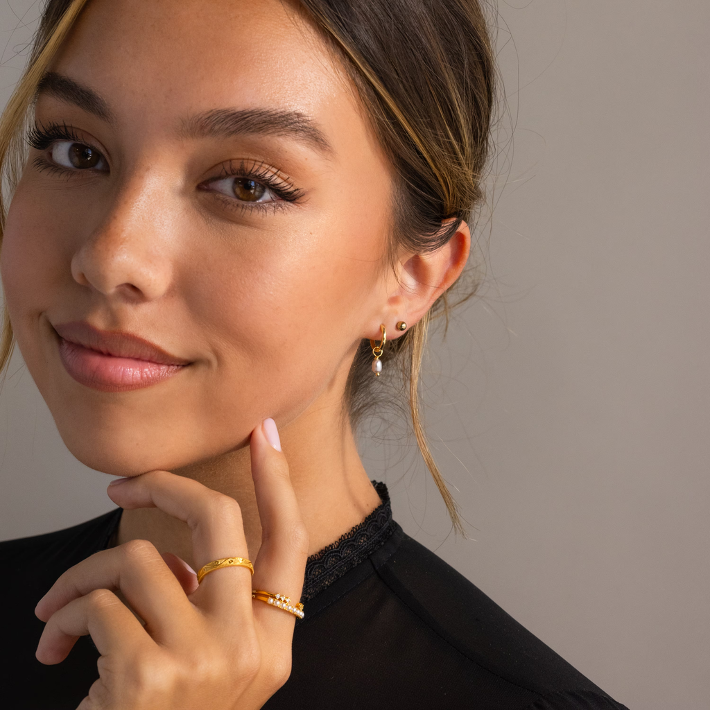 A woman with light brown hair wears gold rings and the Smokey Pearl Drop Huggies, gently resting her hand near her chin while smiling softly.