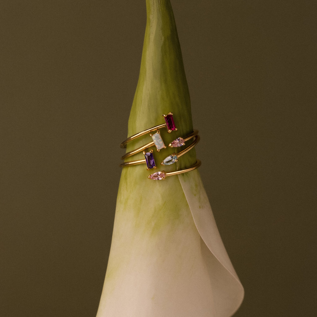 The Toi et Moi Wrap Ring, a gold stacking style with vibrant gemstones, is displayed on the tip of a white calla lily against a brown background.