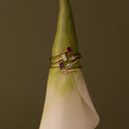 The Toi et Moi Wrap Ring, a gold stacking style with vibrant gemstones, is displayed on the tip of a white calla lily against a brown background.