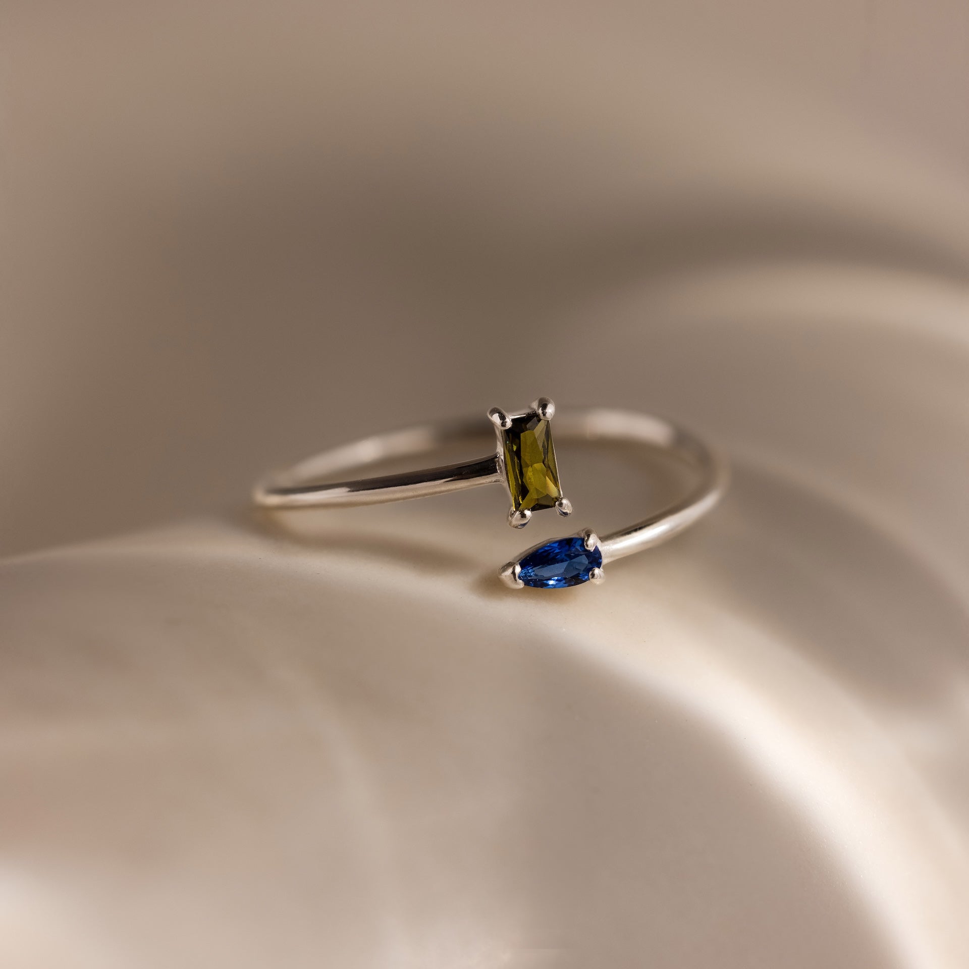 The Toi et Moi Wrap Ring in Silver showcases a rectangular olive gem and a marquise blue gem set on a soft, creamy background for a romantic touch.
