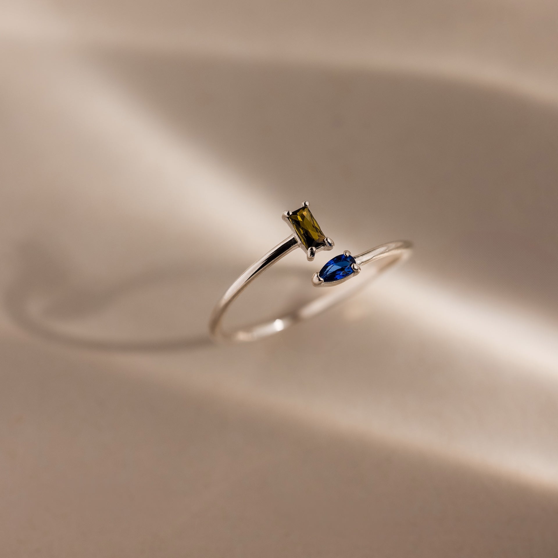 The Toi et Moi Wrap Ring in Silver showcases a rectangular green stone and an oval blue stone, elegantly set together on a sleek, light-toned background.