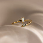 The Toi et Moi Wrap Ring in 18K Gold features a rectangular opal and a teardrop-shaped clear stone, beautifully set against a soft beige background.