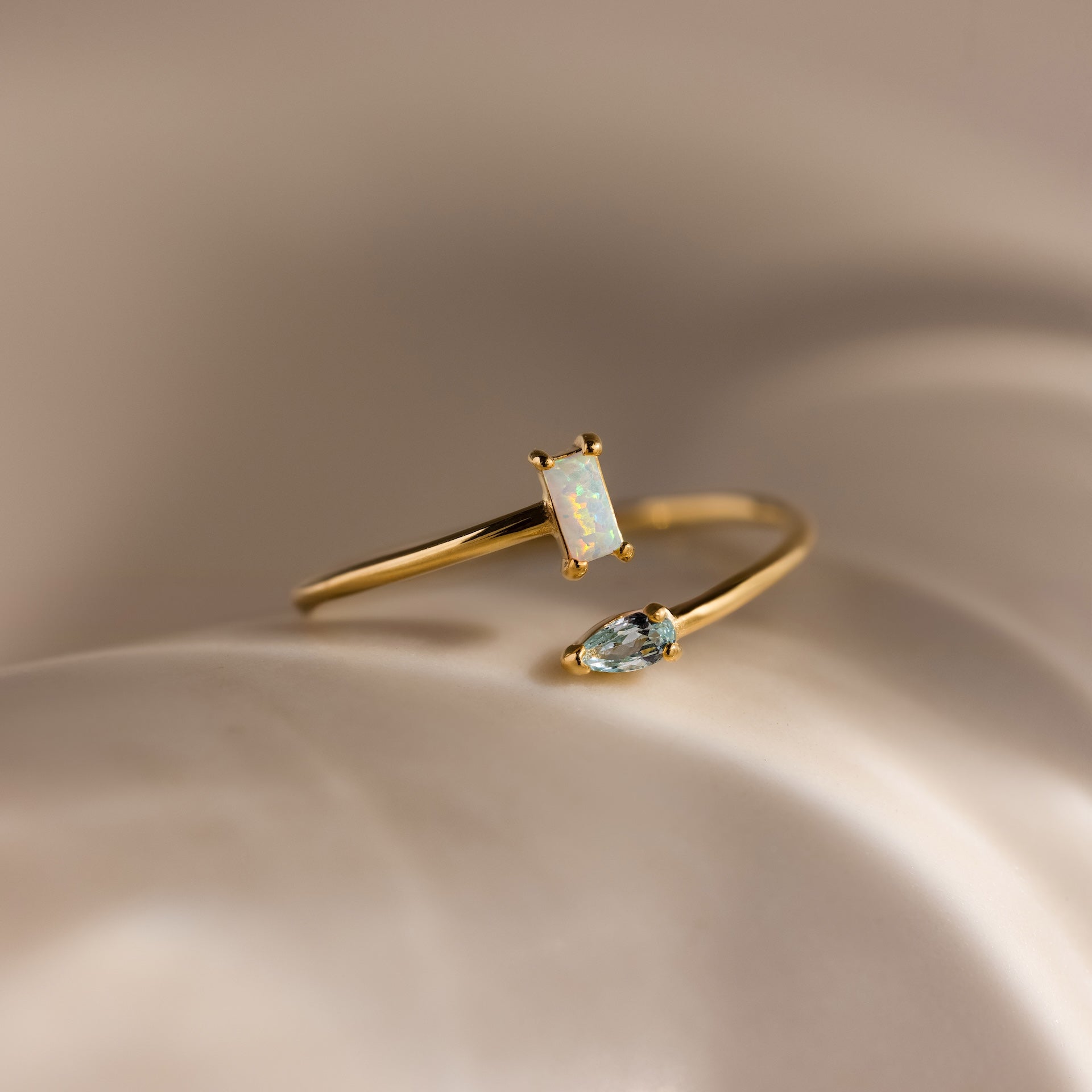 The Toi et Moi Wrap Ring in 18K Gold features a rectangular opal and a teardrop-shaped clear stone, beautifully set against a soft beige background.