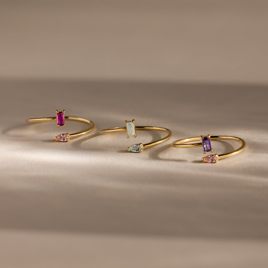 The Toi et Moi Wrap Ring in 18K Gold features delicate overlapping bands adorned with vibrant rectangular and teardrop gemstones, elegantly displayed on a neutral background.