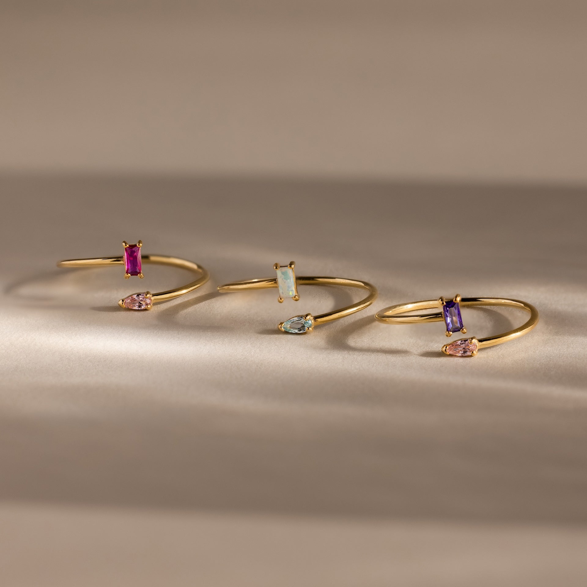 The Toi et Moi Wrap Ring in 18K Gold features delicate overlapping bands adorned with vibrant rectangular and teardrop gemstones, elegantly displayed on a neutral background.