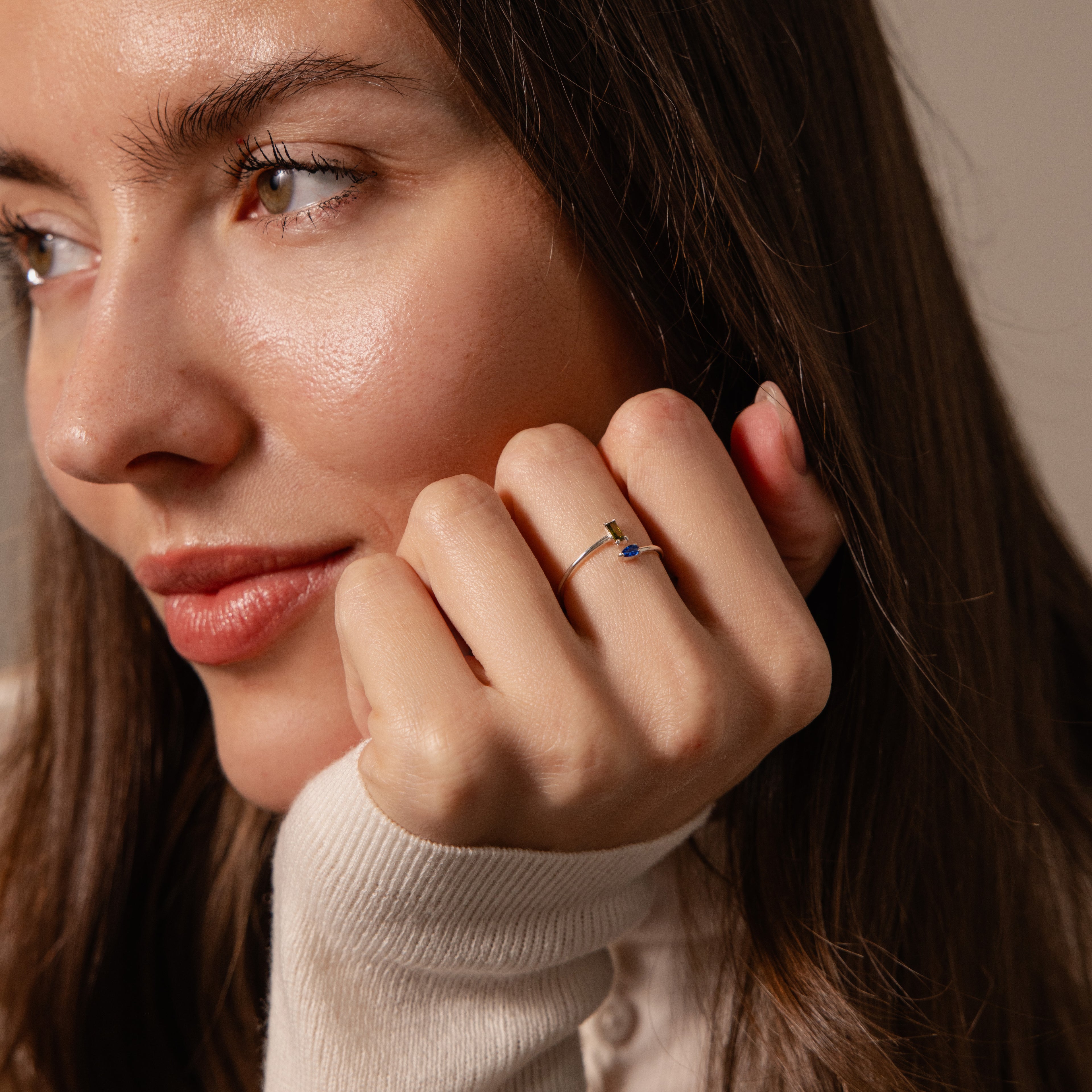 A woman with long brown hair rests her chin on her hand, wearing the Toi et Moi Wrap Ring in gold, featuring a small blue stone.