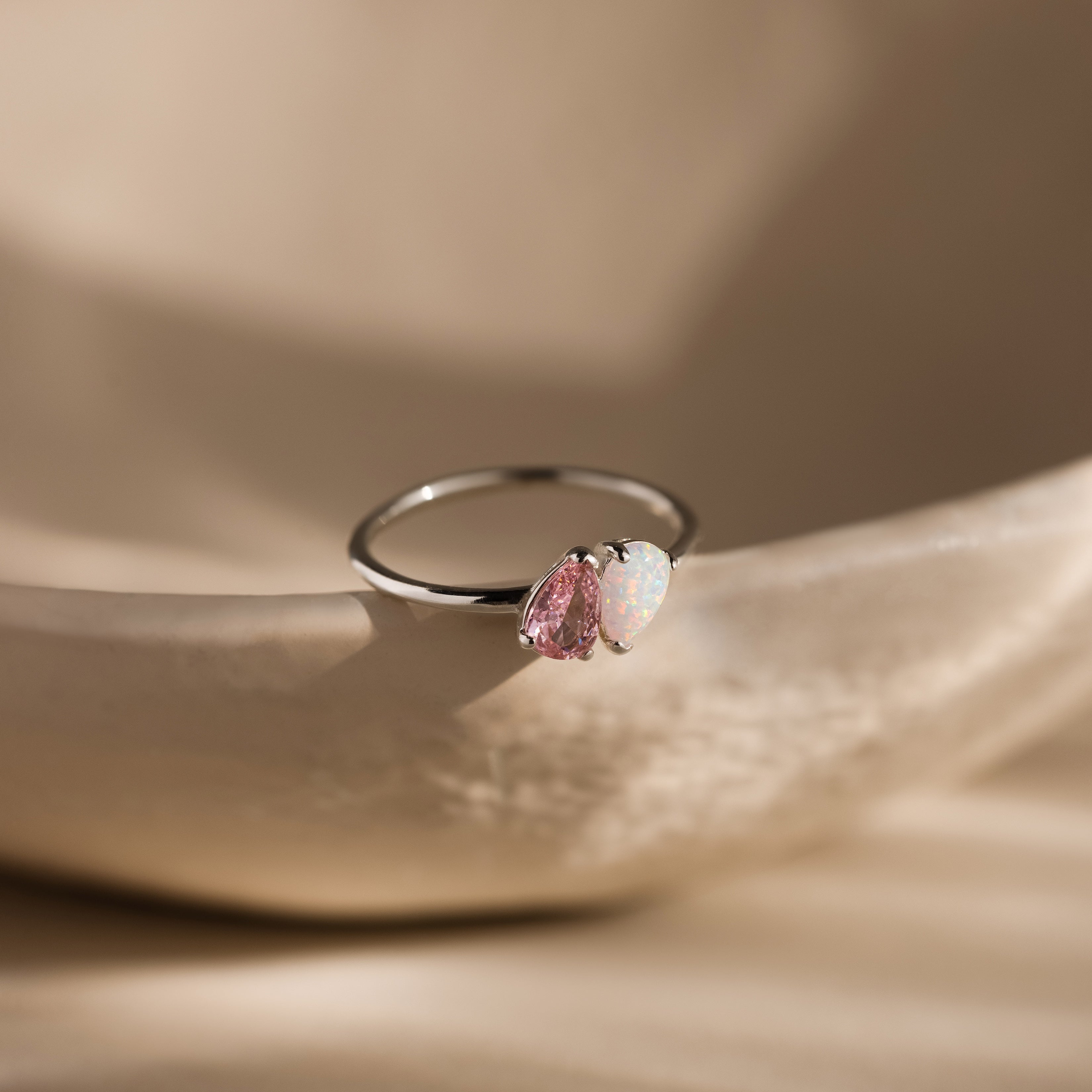 The Duo Pear Toi Et Moi Ring in Silver, featuring pink and white opal pear-shaped stones, is displayed on a smooth beige surface under soft lighting.