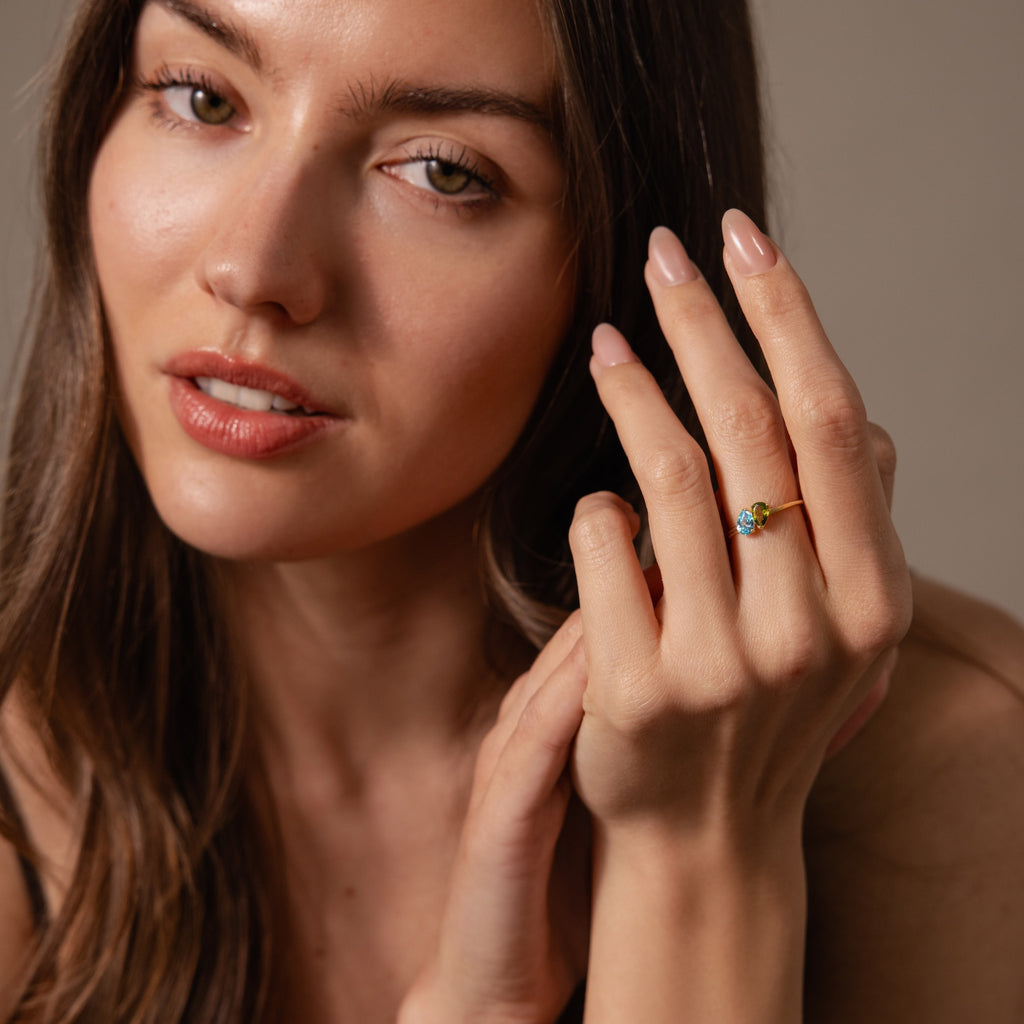 A woman with long brown hair wears the Duo Pear Toi Et Moi Ring in 18K Gold, featuring blue pear-shaped stones, as she gazes softly at the camera.