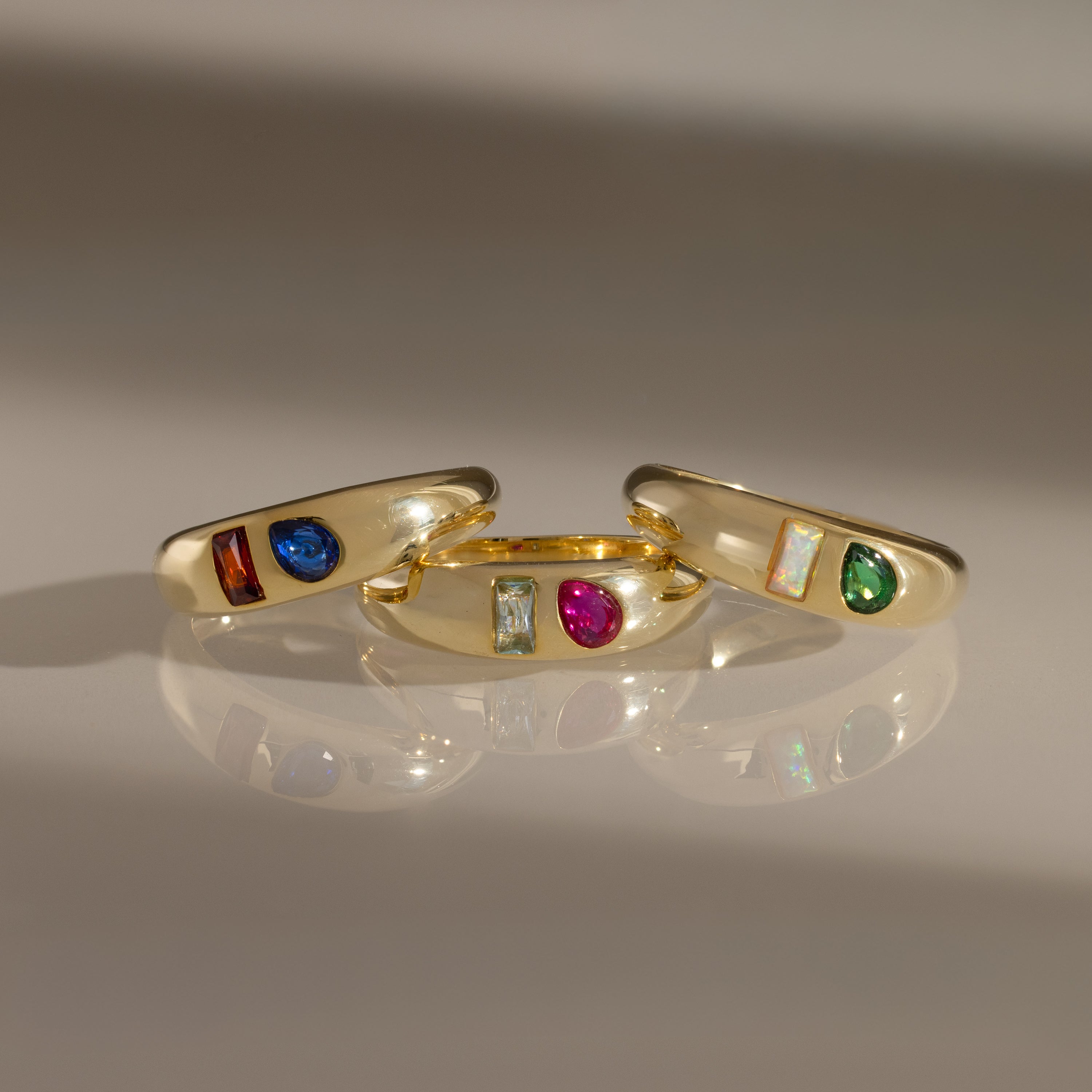 The Toi et Moi Dome Ring in 18K Gold, set with colorful blue, red, green, and iridescent gemstones, sparkles elegantly on a reflective surface.