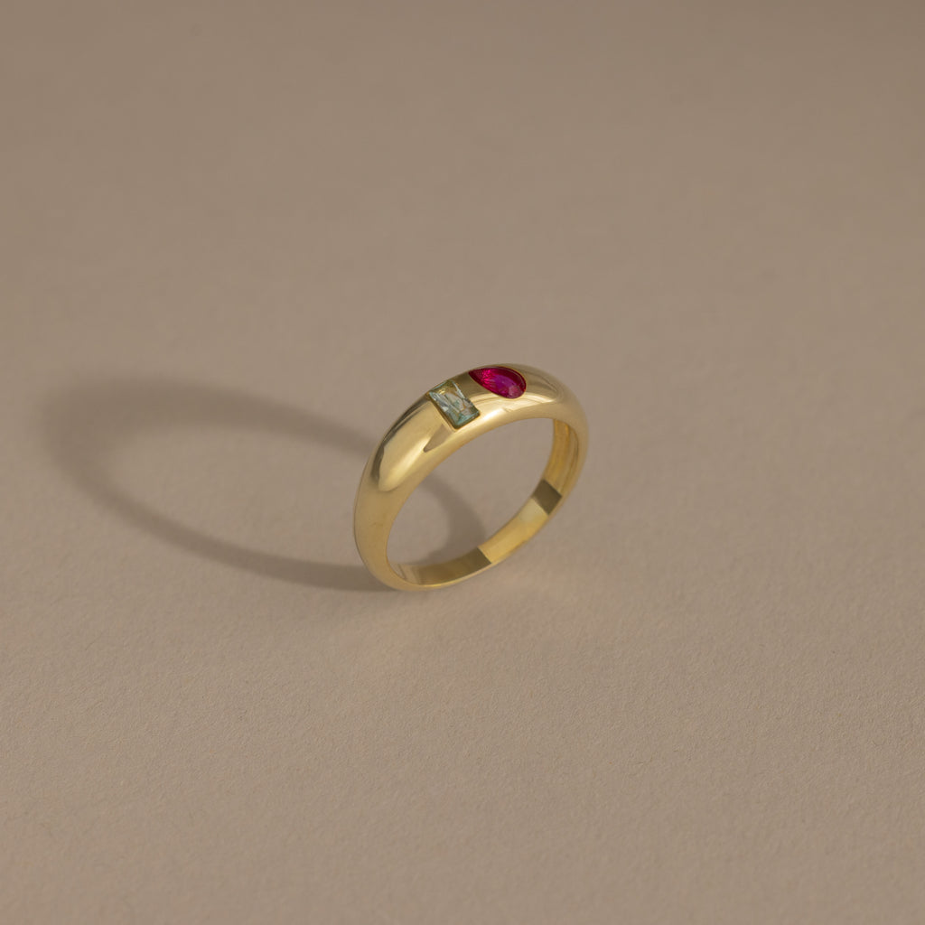 The Toi et Moi Dome Ring in 18K Gold features a rectangular green stone and a round red stone set side by side, displayed on a beige background.