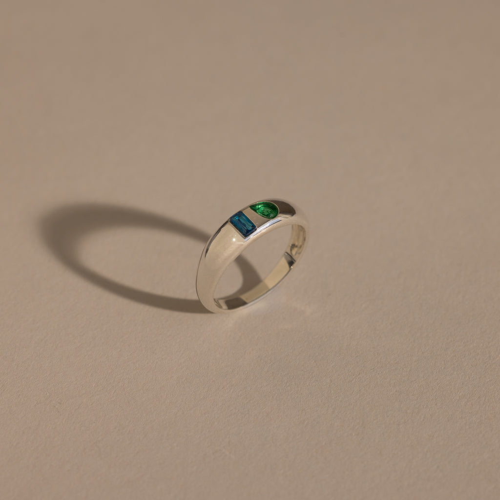 The Toi et Moi Dome Ring in Silver, featuring one blue and one green stone, casts a shadow on a beige surface.