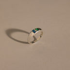 The Toi et Moi Dome Ring in Silver, featuring one blue and one green stone, casts a shadow on a beige surface.