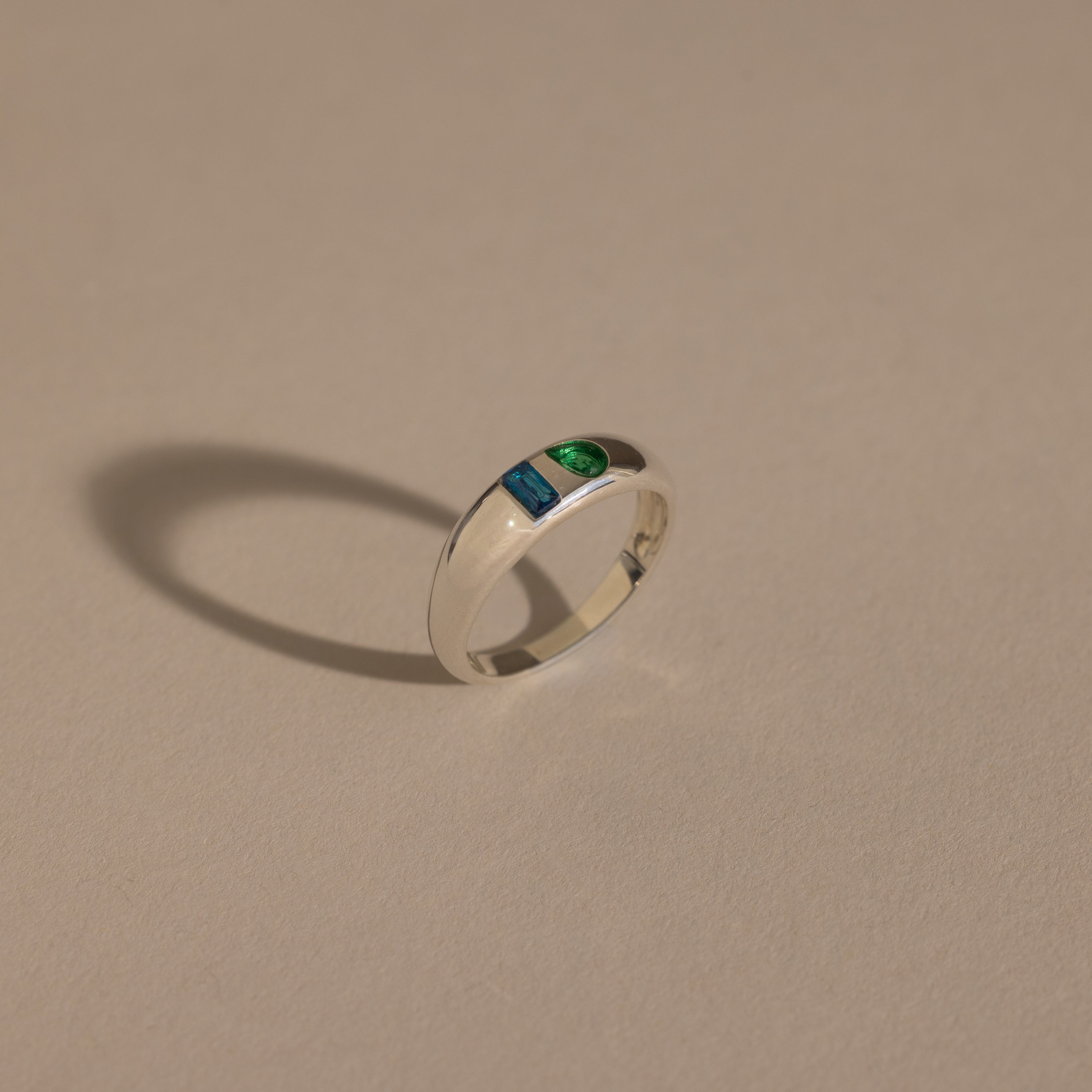 The Toi et Moi Dome Ring in Silver, featuring one blue and one green stone, casts a shadow on a beige surface.