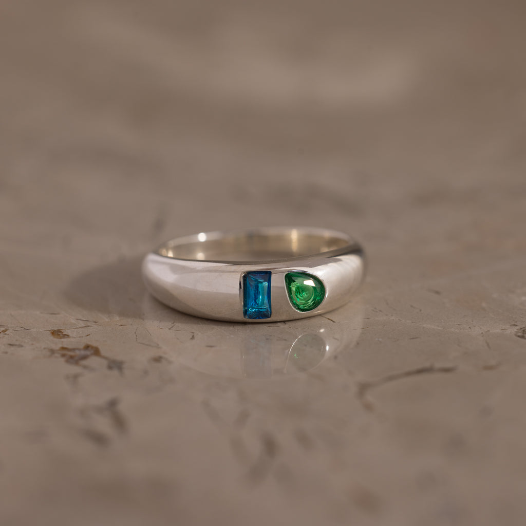 The Toi et Moi Dome Ring in Silver features a blue rectangular gem and a green round gem, displayed on a marble surface.